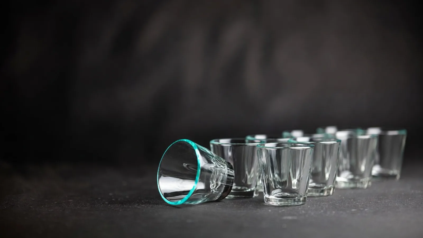 A row of eight small empty drinking glasses on a dark matte surface, one tipped on its side, under clinical studio lighting.