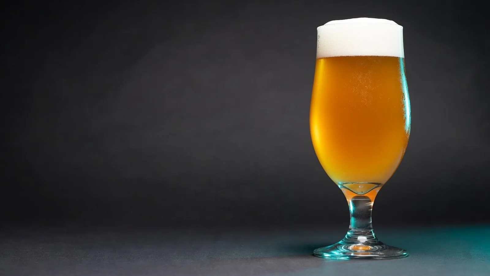 A single plain glass of pale lager on a dark matte surface under clinical studio lighting, with a cyan rim light and faint condensation.