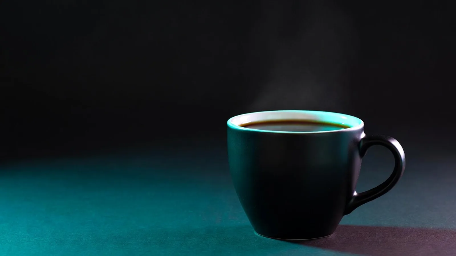 A matte-black cup of black coffee on a dark teal surface, lit with a cyan rim light and faint rising steam.