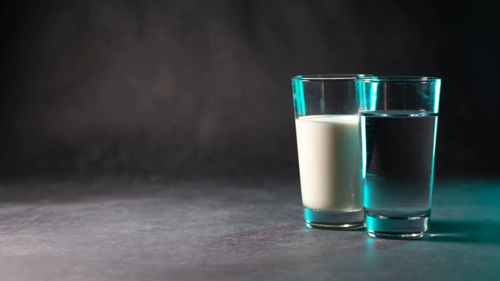 Two clear glasses side by side on a dark matte surface — one of milk, one of water — under clinical studio lighting with a cyan rim light.