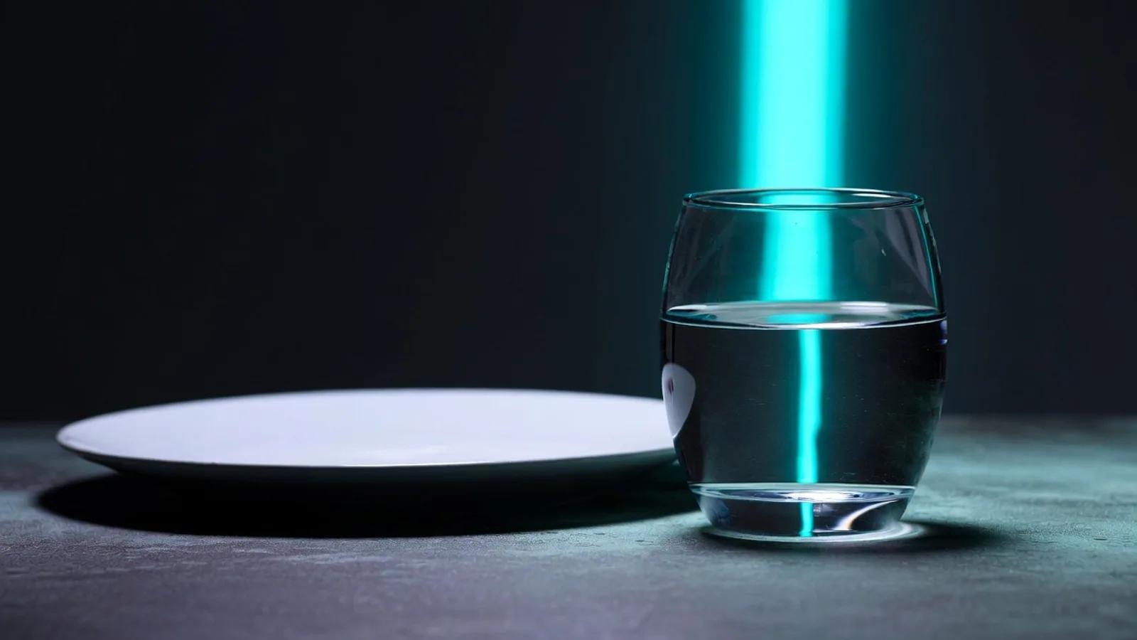 A single clear glass of water beside an empty white plate on a dark matte surface, under clinical studio lighting with a cyan rim light.