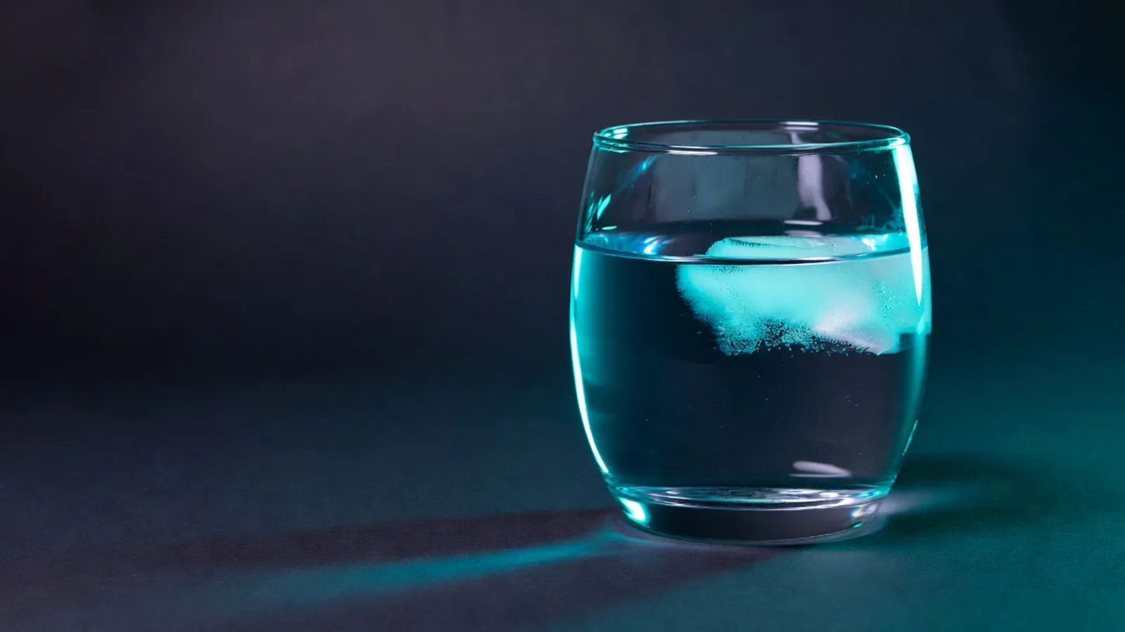 A single clear glass of water beside a smooth matte ceramic surface on a dark base, under clinical studio lighting with a cyan rim light.