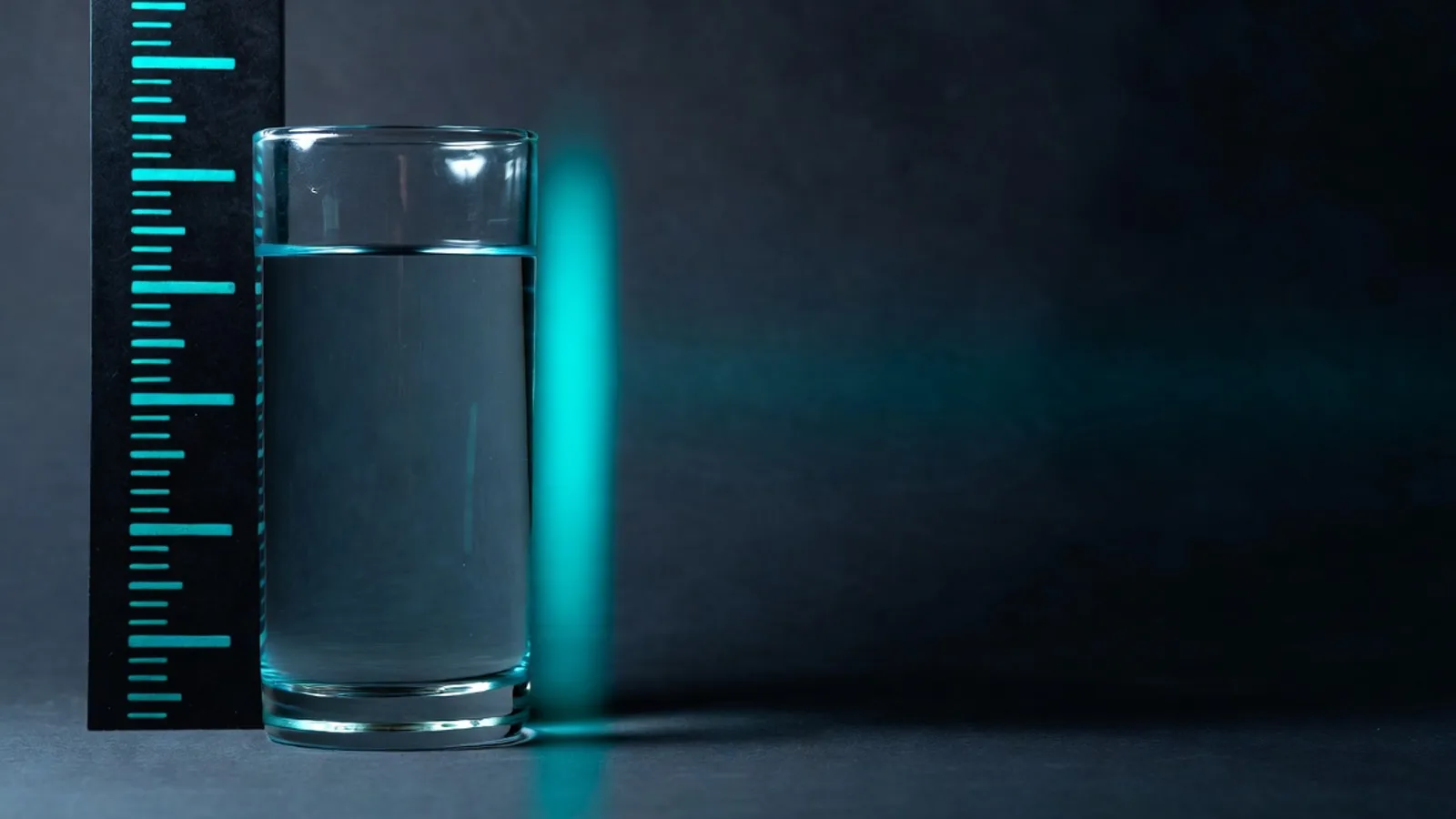 A single tall glass of water on a dark matte surface beside a minimal teal measurement-scale motif, under clinical studio lighting.