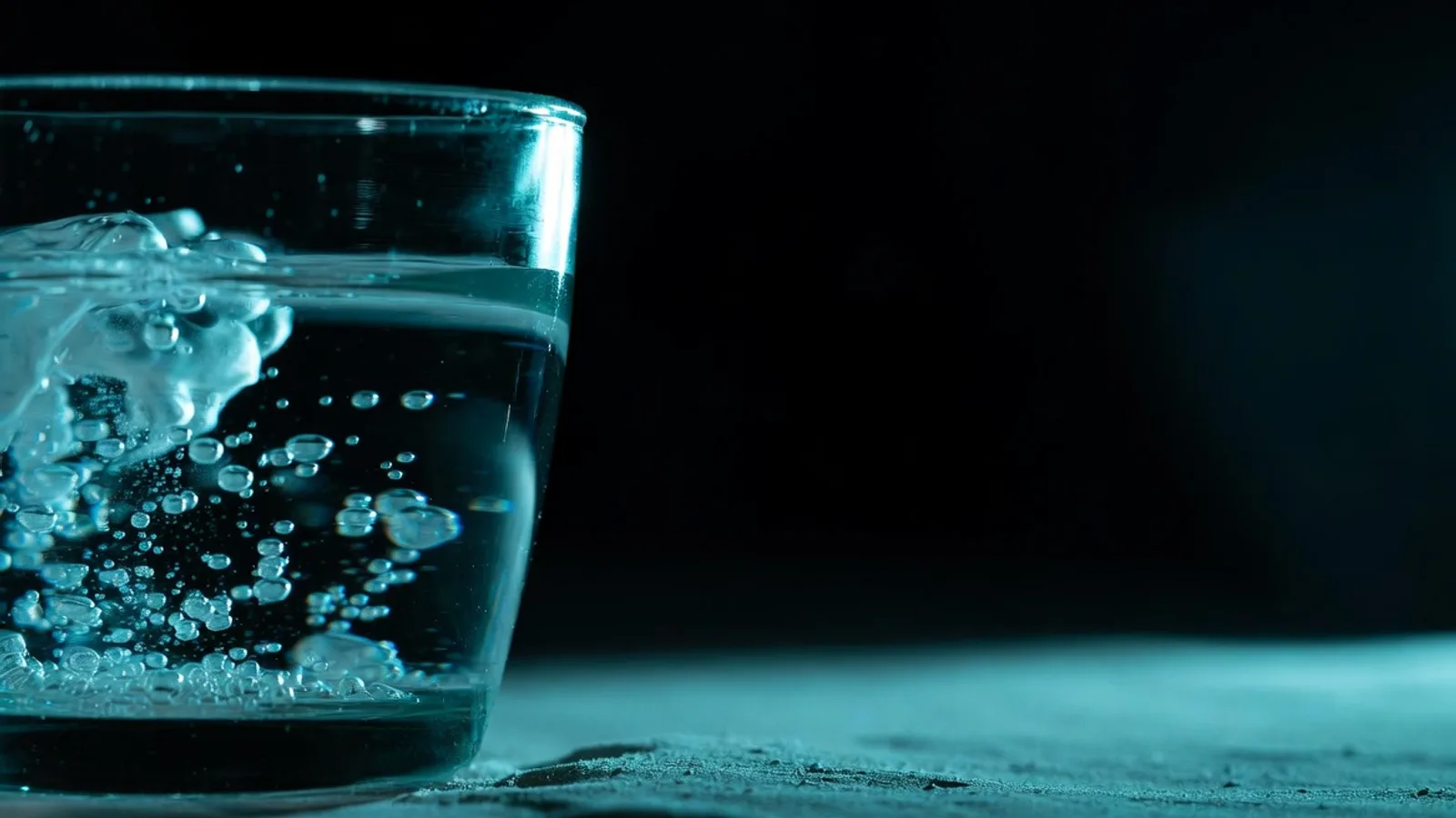 A single clear glass of sparkling water with fine rising bubbles on a dark matte surface, under clinical studio lighting with a cyan rim light.
